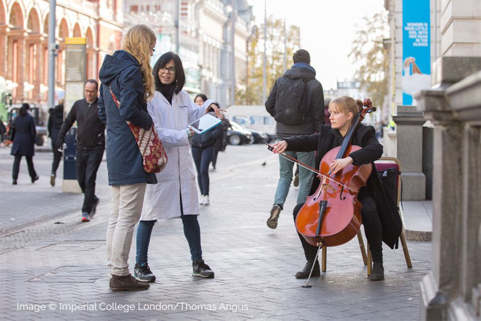 A female student, engaging with a female passer-by, looking at a tablet, with a female cellist, performing on the side walk in front of the Science Museum in South Kensington.